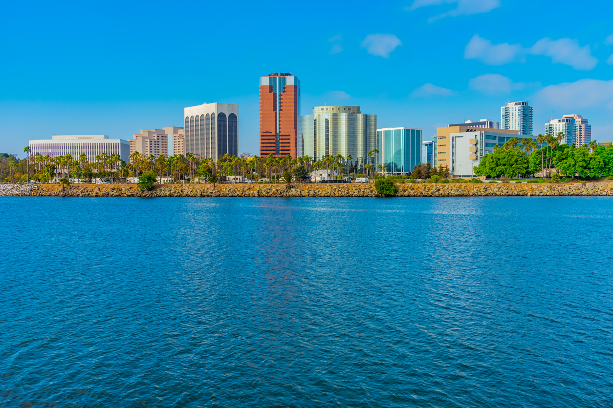 Long Beach skyline stands above the water of the Pacific Ocean. Palm trees and recreational vehicles stand on the edge of the water.